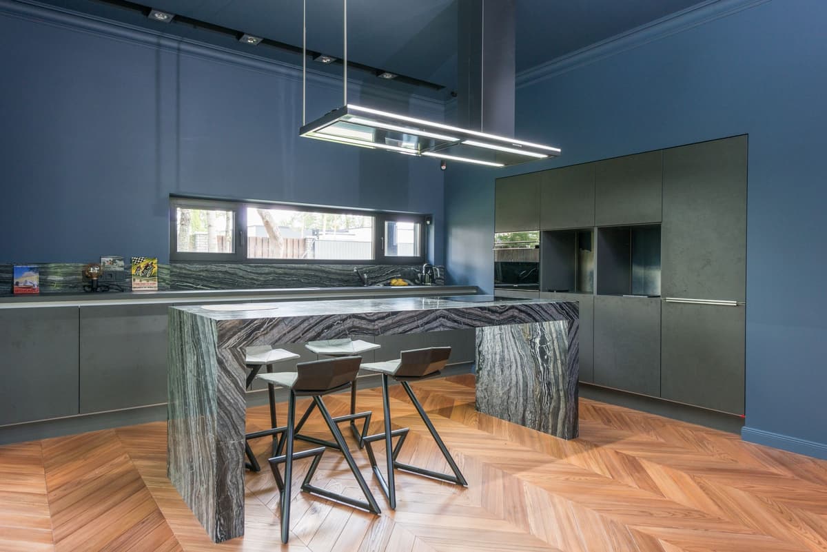 Deep blue kitchen with a dark marble waterfall island, herringbone wood floor and matte dark cabinets